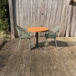 Buitenopstelling met ronde houten horecatafel, twee groene stoelen en een sfeervolle tuinsetting met schutting en zonlicht.