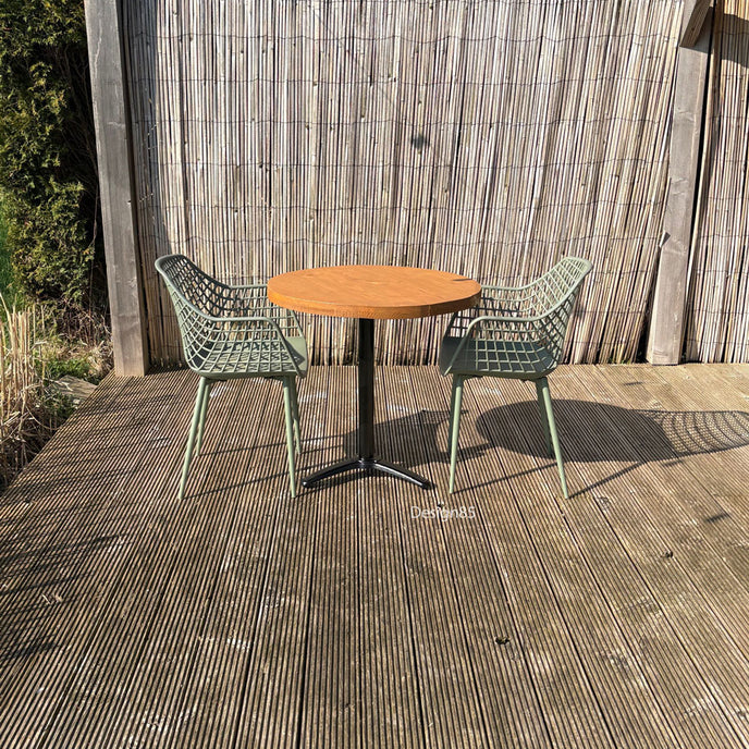 Buitenopstelling met ronde houten horecatafel, twee groene stoelen en een sfeervolle tuinsetting met schutting en zonlicht.