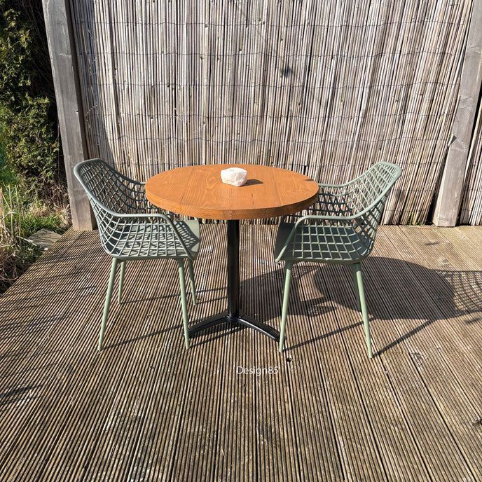 Ronde buitentafel met houten blad en zwart metalen onderstel, gecombineerd met twee groene designstoelen op een houten vlonderterras.