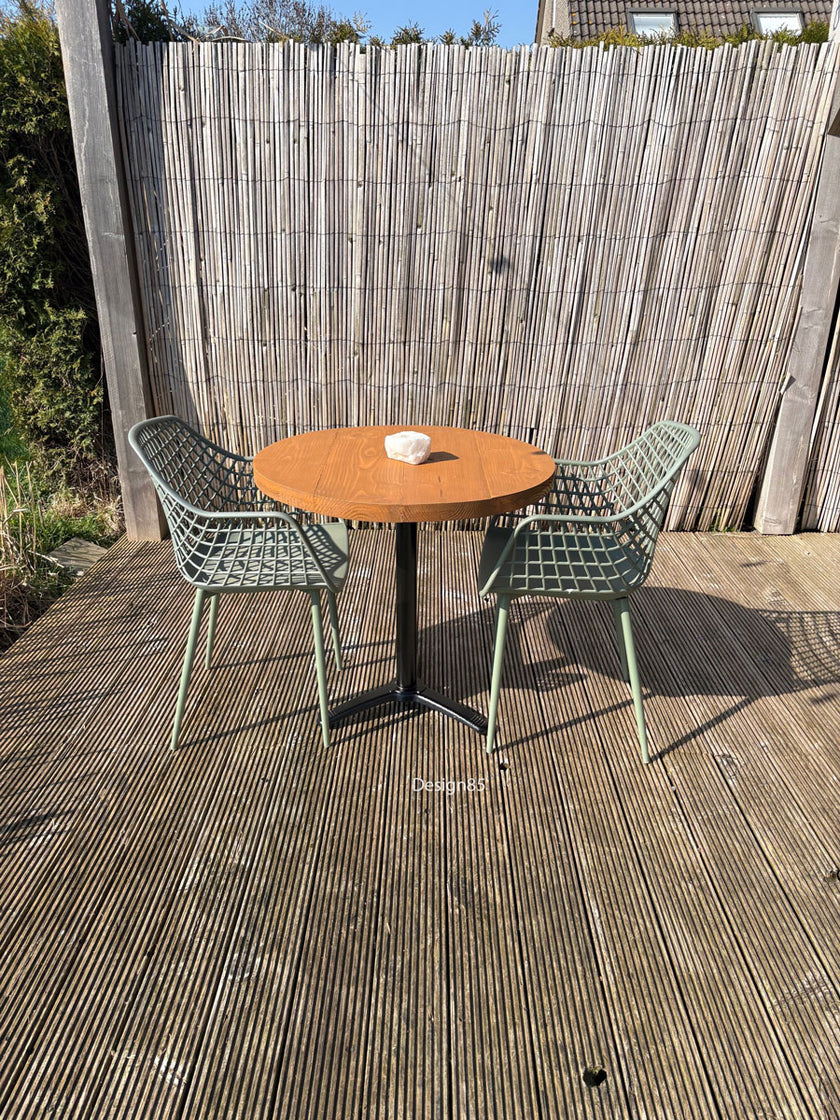 Ronde buitentafel met houten blad en zwart metalen onderstel, gecombineerd met twee groene designstoelen op een houten vlonderterras.