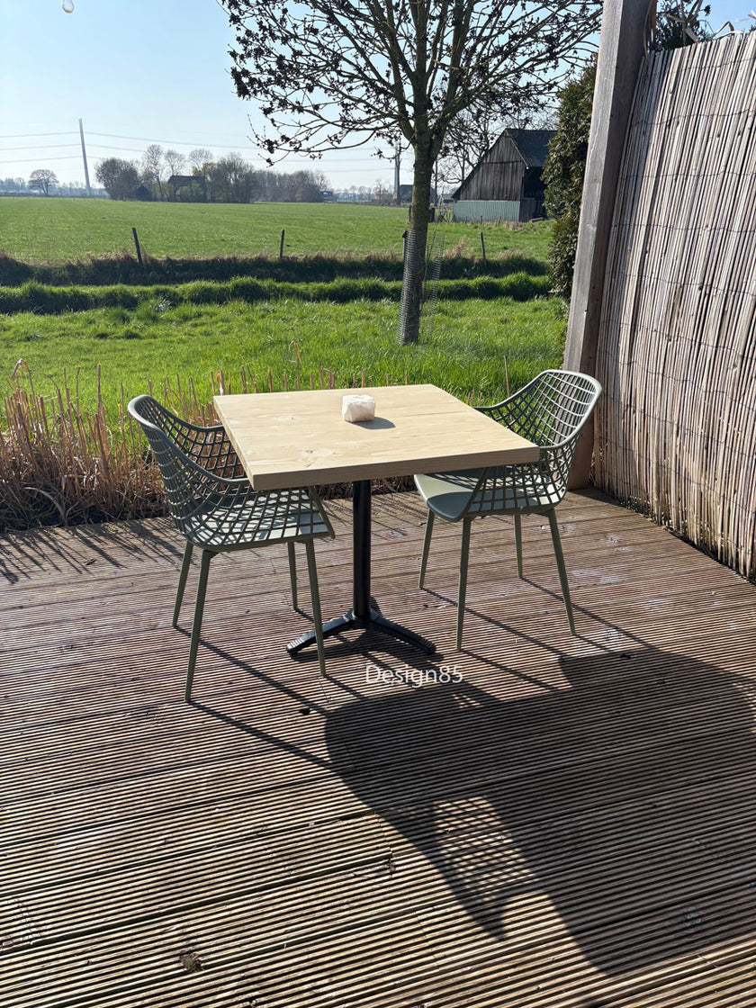 Industriële bartafel met houten blad op een terras in de tuin, omringd door planten en moderne stoelen.