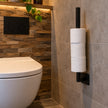 Industriële WC rolhouder Vierkant van zwart staal met drie toiletrollen, aan de muur gemonteerd naast een modern wit toilet in een sfeervolle industriële toiletruimte.