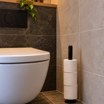 Steigerbuis toiletrol staander Zwart van industrieel staal met drie toiletrollen, vrijstaand naast een modern wit toilet in een sfeervolle industriële toiletruimte.